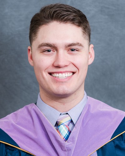 Hunter poses for the camera, smiling. He is wearing a dark blue graduation gown lined with purple, a button-down shirt and colorful necktie.