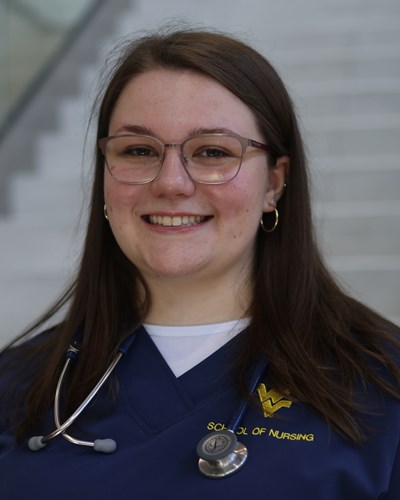 Lindsey smiles while wearing dark blue WVU School of Nursing scrubs and a stethoscope around her neck. Lindsay is seen standing at the bottom of the main staircase in the Pylons lobby of the Health Sciences Center in Morgantown.