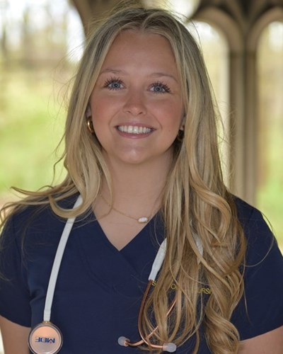 Paige smiles for the camera while wearing dark blue WVU School of Nursing scrubs and a stethoscope draped around her neck. Paige is standing in front of arched openings that reveal a slightly wooded area.
