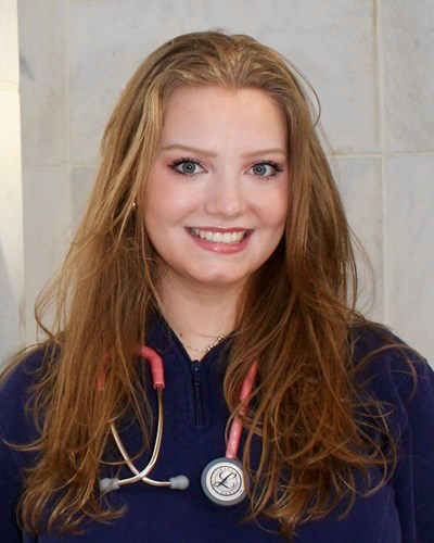 Elizabeth is seen smiling for the camera, wearing a dark blue quarter-zip fleece sweatshirt with a stethoscope draped around her neck. Elizabeth is standing in front of a Pylon in the entrance to the Health Sciences Center in Morgantown.