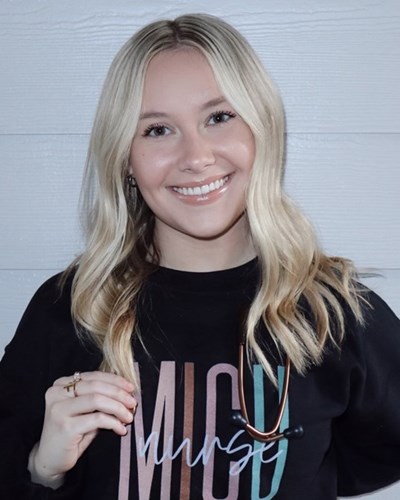 Abigail is smiling, standing in front of a white-painted wood panelled wall, and wearing a “MICU nurse” sweatshirt with a stethoscope draped around her neck.