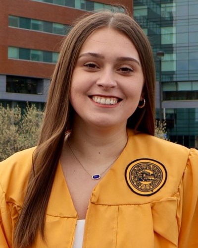 Mallori smiles as she is seen wearing a gold graduation gown, and is standing in front of WVU Medicine’s J.W. Ruby Memorial Hospital in Morgantown.