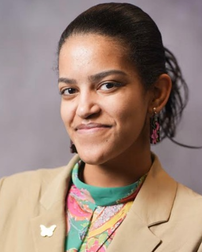Yohanna poses for a portrait, wearing a colorful teal and pink blouse with a tan sportcoat.