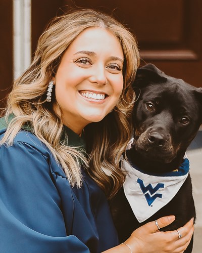 Anna is sitting on the steps in front of a WVU classroom building, wearing a blue grauation gown. Anna has a dark brown dog that’s wearing a white bandana with a blue “Flying WV” logo on it around its neck sitting next to her