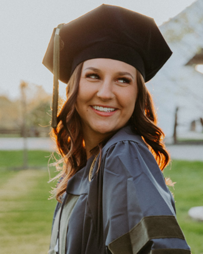 Kaleigh stands in an open grassy area turned away from the camera and looking over her left shoulder. She is smiling while wearing a cap and gown