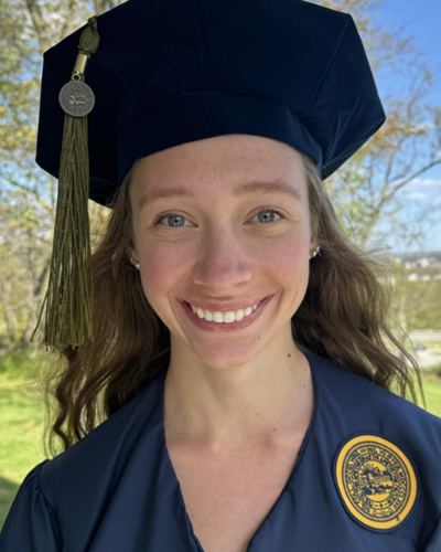 Emily smiles, wearing a dark blue cap and gown, while in an outdoor setting.