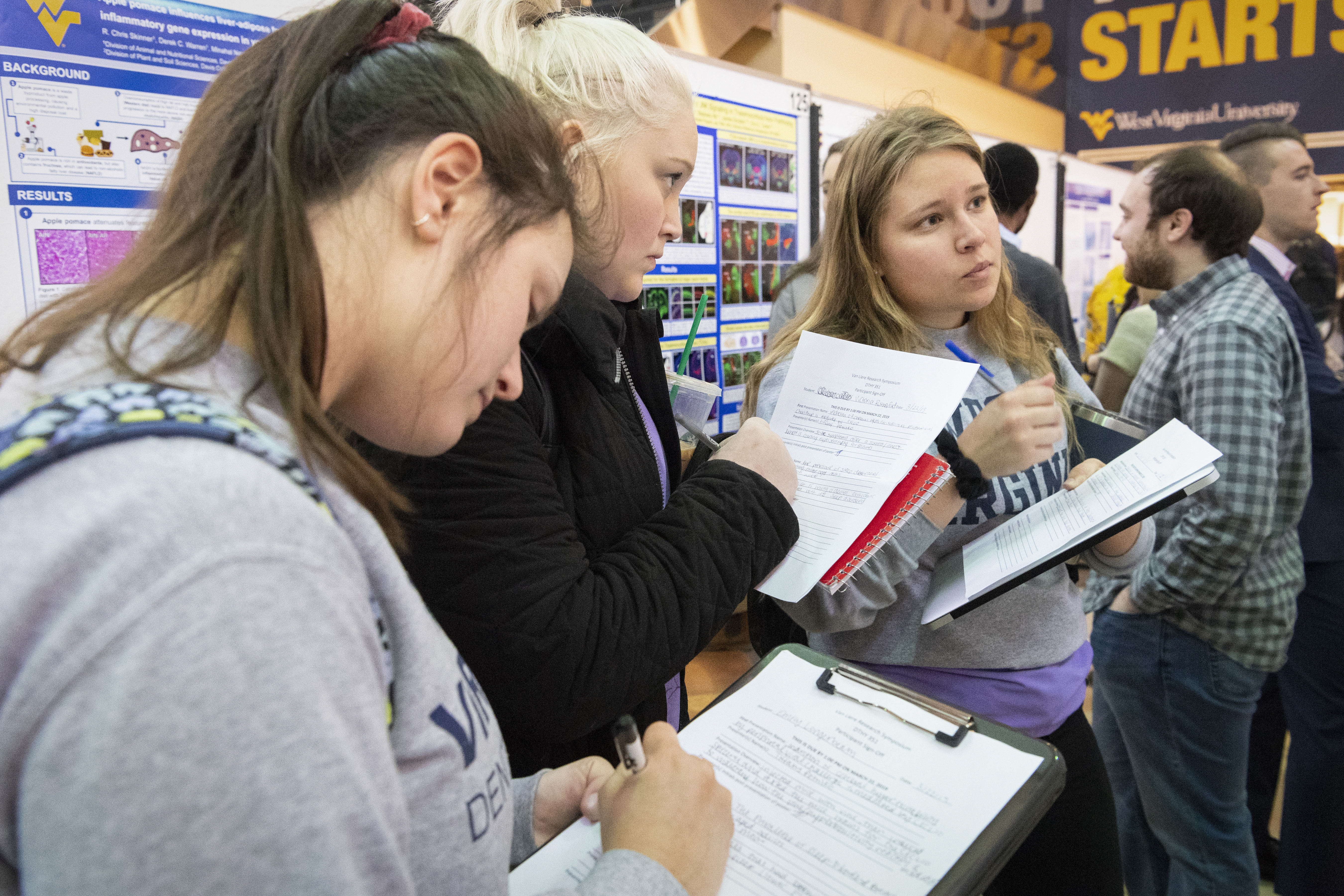 A group of three student attendees stand amongst the poster presentations of the Pylons area if the Health Sciences Center. Each is holding a clipboard and are observing and note-taking.