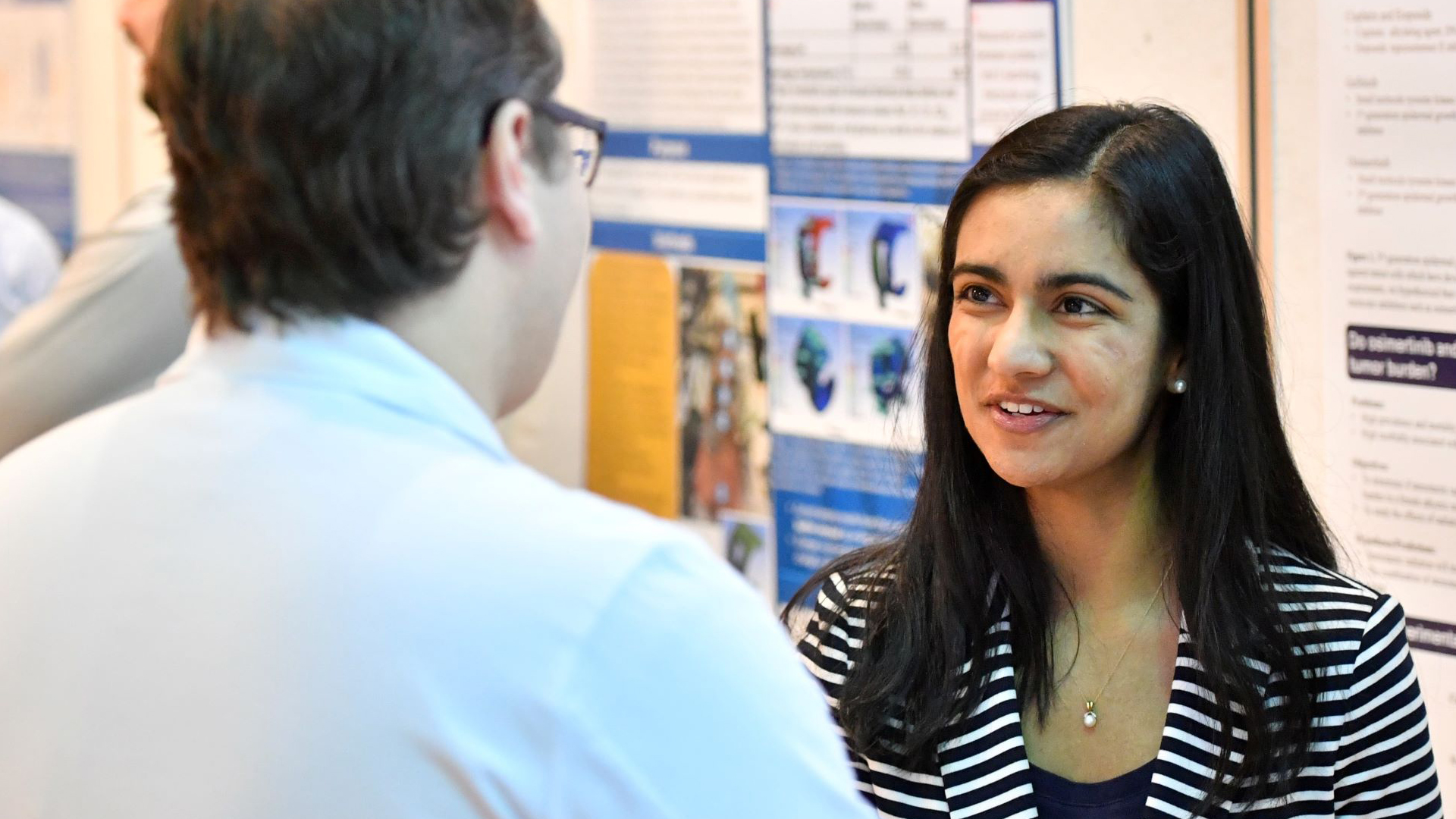 A student, shown to the right, is in discussion with another individual who is to the left and facing away. They are standing in front of a series of poster presentations inside the Pylons area of the Health Sciences Center.