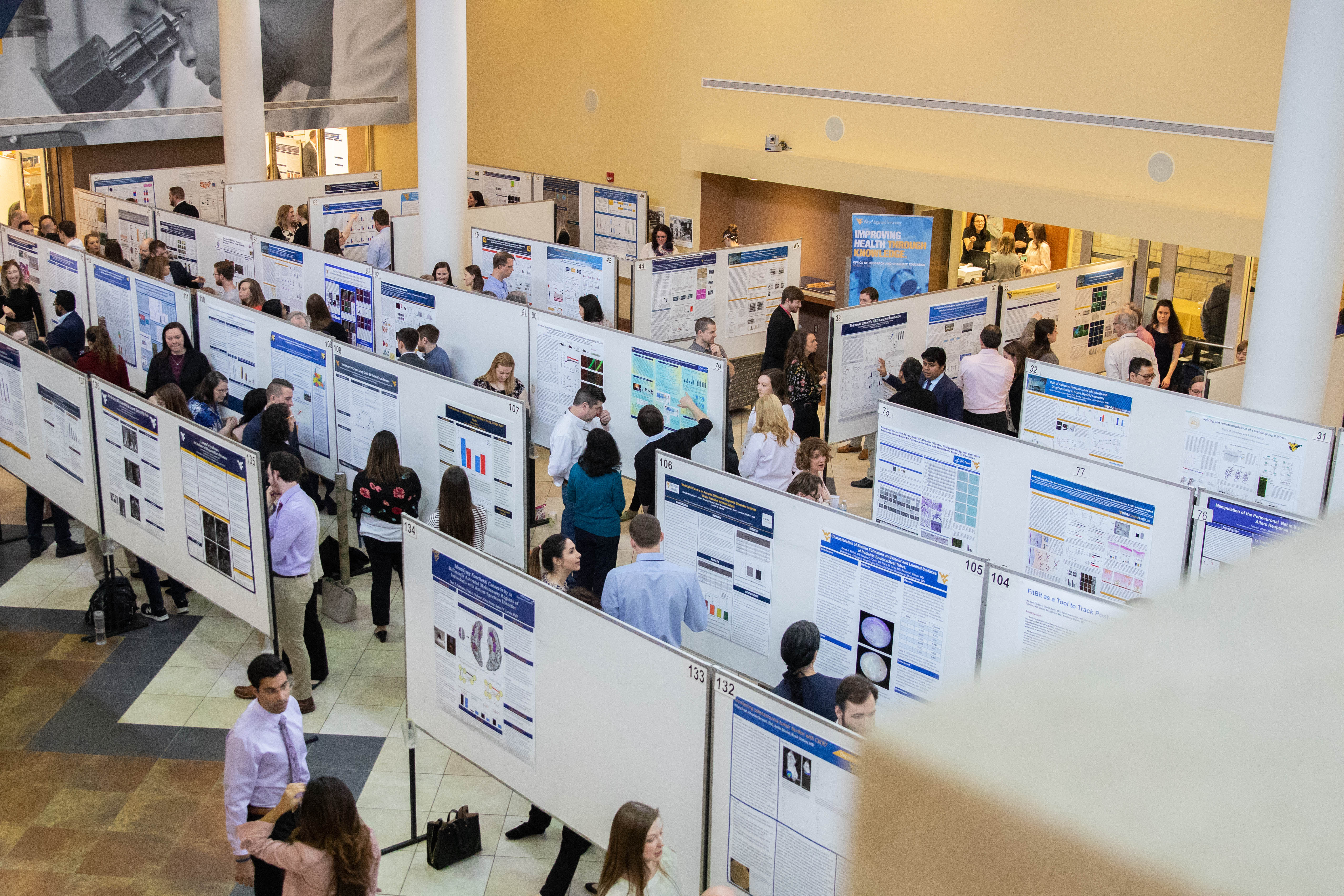 A large number of people are seen mingling amongst several rows of poster displays that are set up in the Pylons area of the Health Sciences Center in Morgantown. Some people are studying the posters, while others are in conversation about the displays.