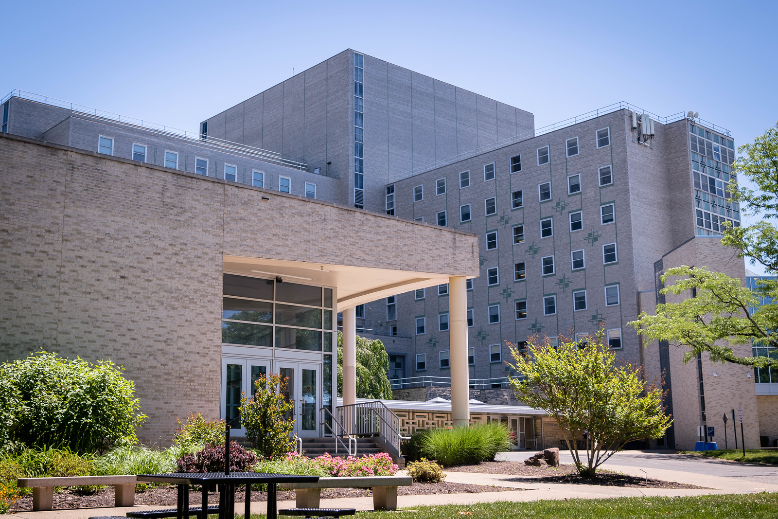 The front exterior of the Health Sciences Center complex show. The mostly white building has several benches and picnic tables on its lawn area, which also has plenty of plant life.