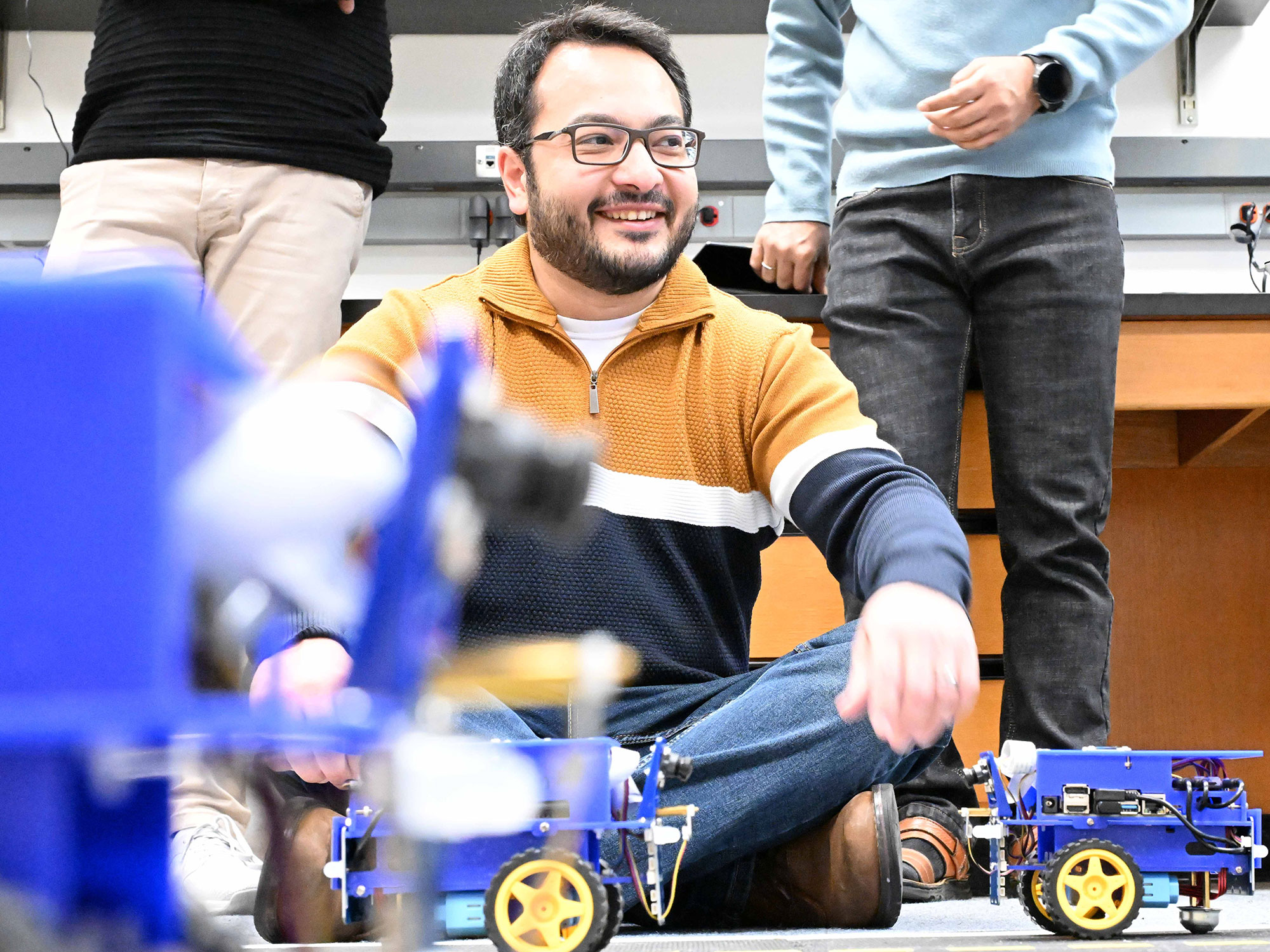 The F1Tenth robot is placed on the floor with a fellow researcher sitting nearby.