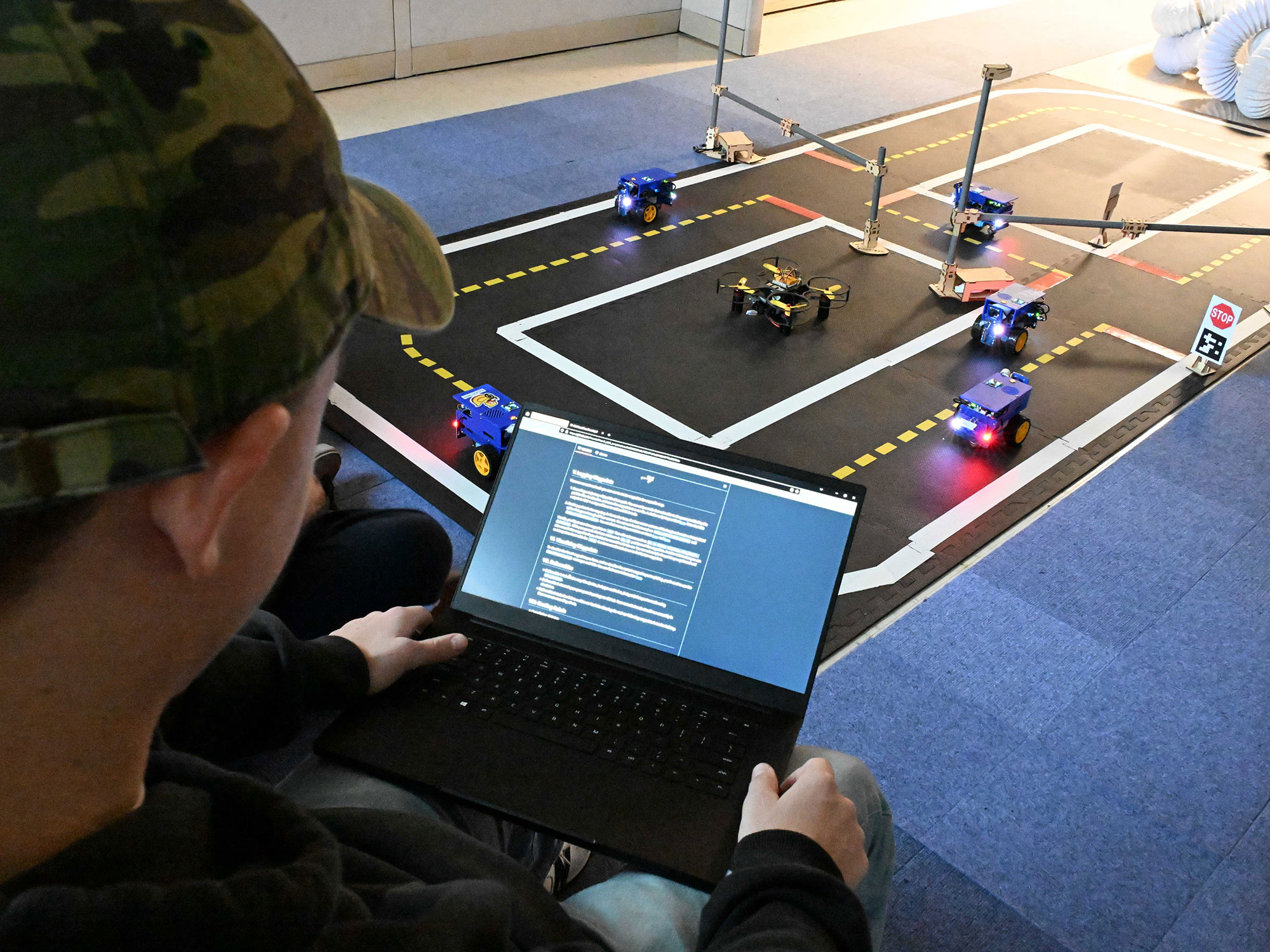 The F1Tenth robot is on a miniature track with marked traffic lanes and some obstacles.  A research on a computer is in the foreground getting ready to run some code for the robot in the simulated environment.. 